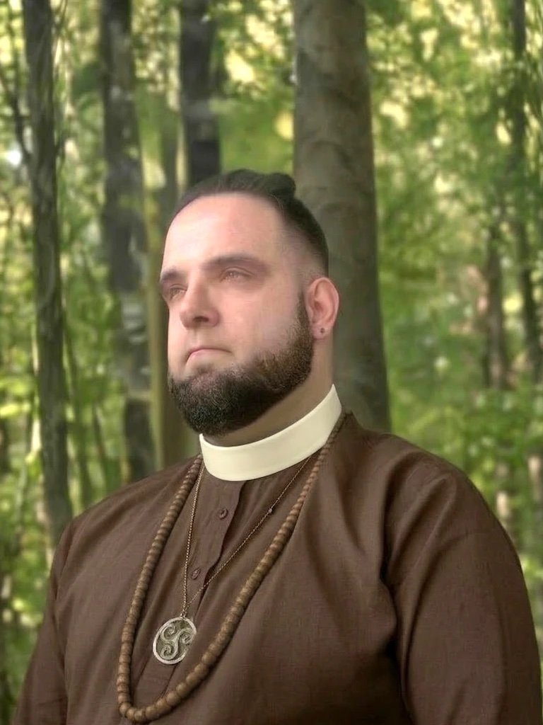 Casey Das in a clerical collar, brown robe, and wooden prayer beads with a triskele pendant, standing in a green forest